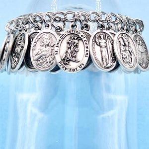 May include: A silver-toned charm bracelet featuring oval pendants with religious figures and inscriptions. The bracelet is suspended in front of a clear glass object, with a light blue background. The charms are connected by a chain.