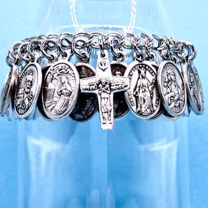 May include: A silver-toned bracelet featuring oval religious medallions and a central cross pendant. The medallions depict various saints and religious figures. The bracelet is displayed against a light blue background.