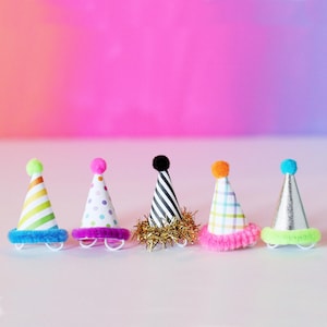 May include: Five miniature party hats arranged in a row. The first has green and yellow stripes with a blue fuzzy trim. The second has colorful polka dots and a purple fuzzy trim. The third has black and white stripes with gold tinsel. The fourth has a plaid pattern and pink fuzzy trim. The fifth is silver with a neon green fuzzy trim.