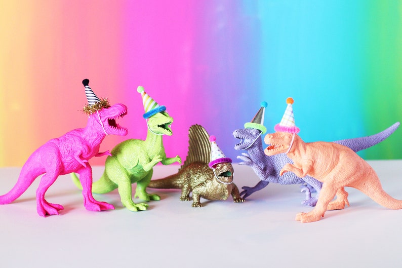 May include: Five colorful plastic dinosaur toys wearing party hats. The dinosaurs are pink, green, gold, purple, and orange.
