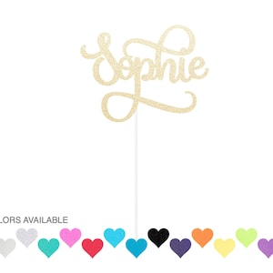 May include: A gold glitter cake topper with the name "Sophie" in a cursive font. The topper is attached to a white stick. The bottom of the image shows a selection of heart-shaped color options, with the text "15 COLORS AVAILABLE".