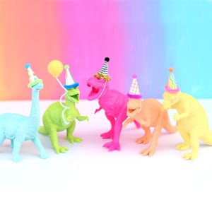 May include: Five plastic dinosaur figurines in various colors including light blue, green, hot pink, peach, and yellow. Each dinosaur wears a small, colorful party hat, and the green dinosaur holds a yellow balloon on a white string.