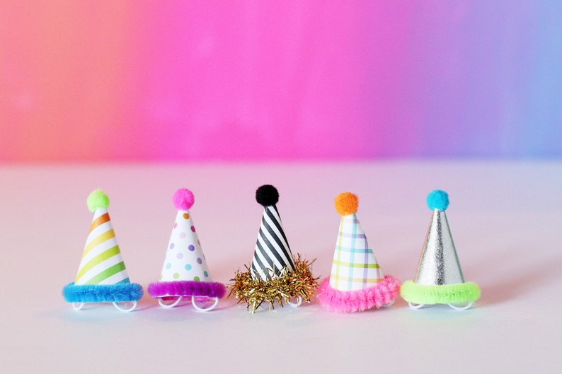May include: Six colorful party hats with different patterns and pom-poms on top. The hats are arranged in a row on a white surface.