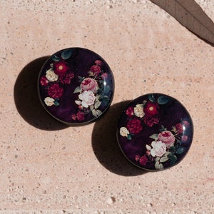 May include: A pair of round, black earrings with a floral design. The earrings feature a dark purple background with pink, white, and red flowers.