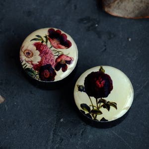 May include: Two round, black, wooden ear gauges with floral designs. One gauge features a dark red rose with green leaves. The other gauge features a cluster of pink and red flowers with green leaves.