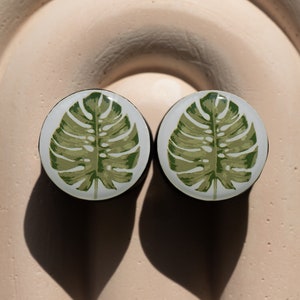 May include: Two round, white, decorative plugs with a green tropical leaf design.