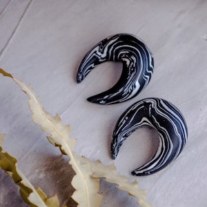 May include: Two black and white marbled crescent shaped ear gauges. The gauges are made of a smooth, polished material and have a swirling pattern.