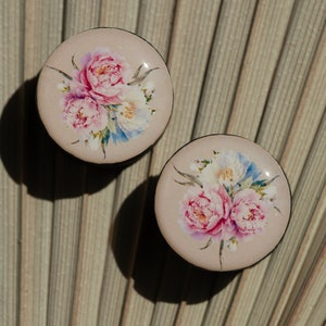 May include: Two round, cream-colored earrings with a floral design. The flowers are pink, white, and blue.