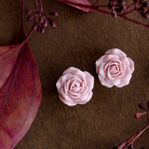 May include: Two pink rose-shaped polymer clay earrings on a brown wooden surface with red leaves.