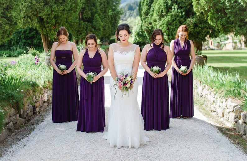 May include: A group of five women in a wedding party. The bride is in the center wearing a white lace wedding dress and holding a bouquet of flowers. The bridesmaids are wearing long, purple dresses and holding small bouquets of white flowers.