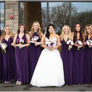 May include: A group of bridesmaids in purple dresses stand with the bride in a white wedding dress. They are all holding bouquets of flowers.