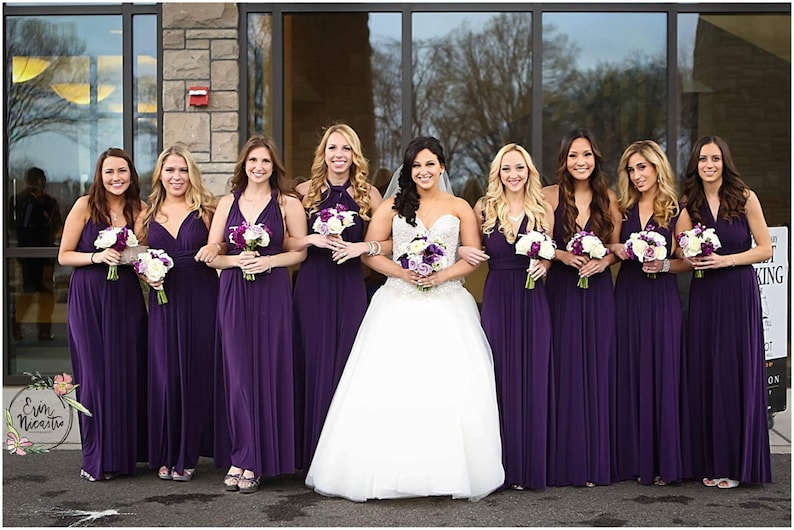 May include: A group of bridesmaids in purple dresses stand with the bride in a white wedding dress. They are all holding bouquets of flowers.