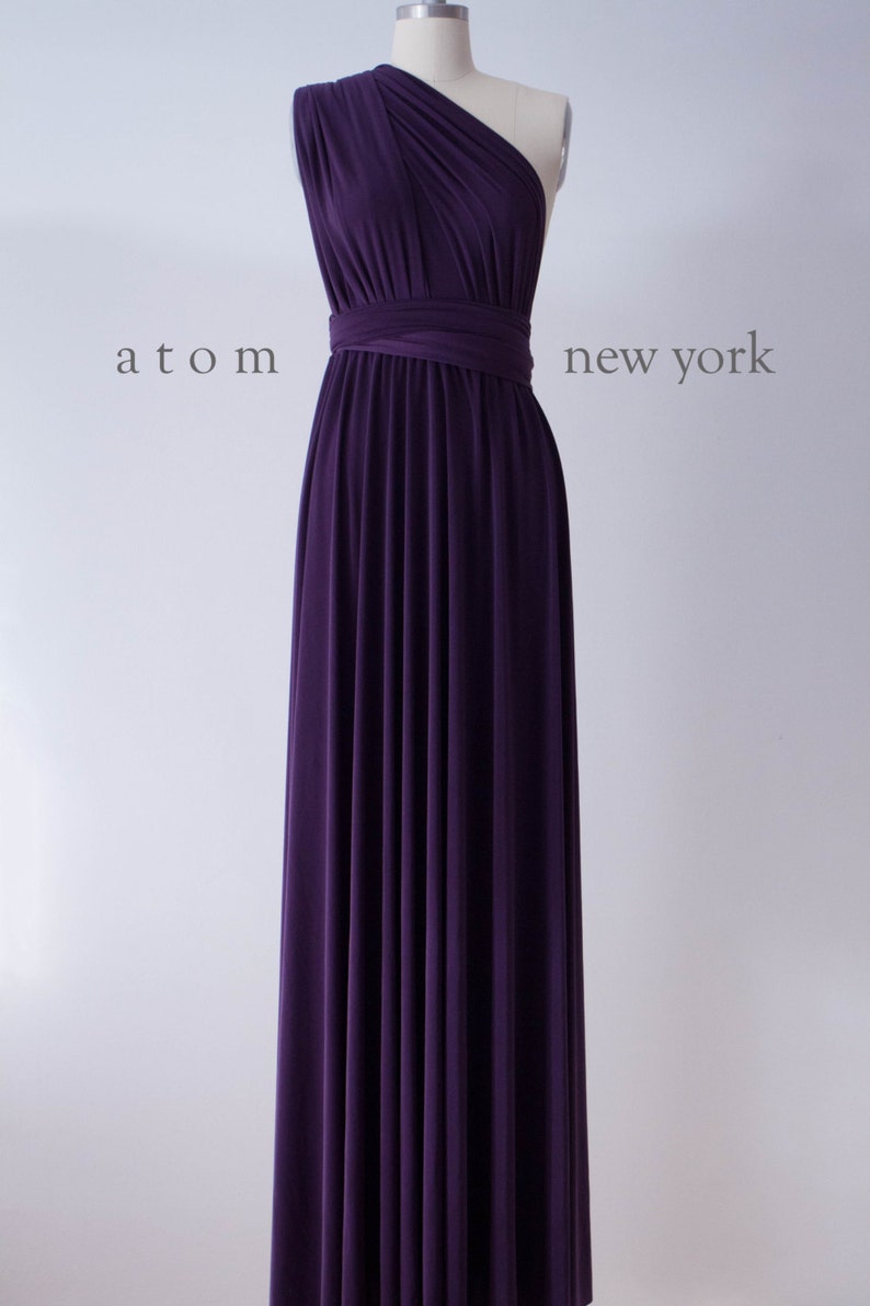 May include: A long, purple, one-shoulder dress with a wrap style bodice. The dress is made of a soft, flowing fabric and has a simple, elegant design. The dress is shown on a mannequin against a white background. atom new york