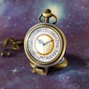May include: A gold-toned pocket watch with a round face and a chain. The watch face has Roman numerals and a design of symbols around the edge.