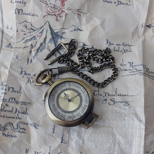Pu&ograve; includere: Un orologio da tasca dorato con una catena, che presenta una mappa della Terra di Mezzo de Il Signore degli Anelli. Il quadrante dell'orologio ha la scritta "Colazione, 2a colazione, Pranzo, Merenda, Cena, Cena".