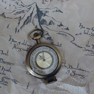 Pu&ograve; includere: Un orologio da tasca in stile vintage con finitura dorata e quadrante nero. Il quadrante dell'orologio ha indici orari con numeri romani e una lancetta nera. L'orologio &egrave; su una catena e si trova su una mappa con un drago e montagne.
