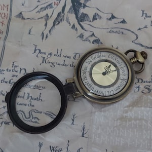 Pu&ograve; includere: Un orologio da tasca in stile vintage con una lente d'ingrandimento. Il quadrante dell'orologio ha numeri romani e le parole "2nd Breakfast, Elevenses, Lunch, Afternoon, Supper, Breakfast". L'orologio &egrave; appoggiato su una mappa con le parole "Here of old was the King under the" e "West lies Greenwood the Great".