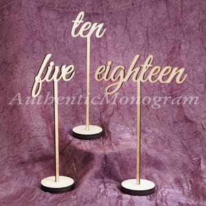 May include: Three wooden table number signs with the numbers "five", "ten", and "eighteen" written in a cursive font. The signs are on wooden stands.