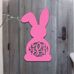 May include: Pink wooden bunny rabbit cutout with a monogrammed circle in the centre. The monogram is the letters "G P B".