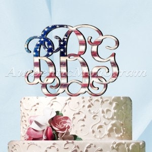 May include: A wooden cake topper featuring the letters "B", "R", and "G" in a decorative script, painted with an American flag design. The topper is placed on a white tiered cake with floral decorations.