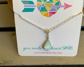 May include: A silver chain necklace with a light blue teardrop-shaped pendant. The pendant is made of wire wrapped around a stone. The necklace is on a white card with the text "you make my heart smile" and a website address.