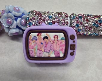 May include: A purple retro television charm with a group of people in pink outfits on the screen. The charm is surrounded by a beaded bracelet with pink, blue, and clear rhinestones and light blue flower beads.