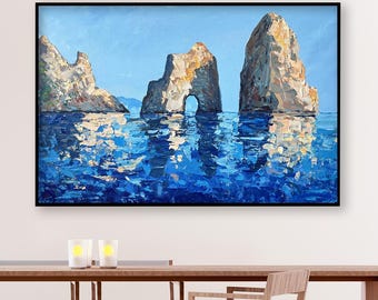 May include: An oil painting depicts three large rock formations in the sea under a blue sky. The water reflects the sky and the rocks. The painting is framed in black and hangs above a wooden table with chairs.