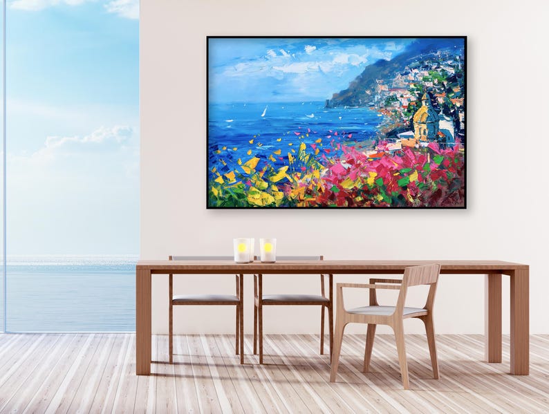 Original hand-painted oil painting on canvas depicting Positano on the Amalfi Coast, with colorful cliffside houses, deep blue sea, and vibrant Mediterranean flowers, framed and hanging on a bright dining room wall above a wooden table with sea view.