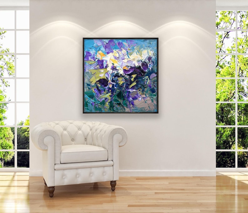 Square original oil painting with bold abstract purple and blue flowers, hanging above a white armchair near a large window in a modern living space.