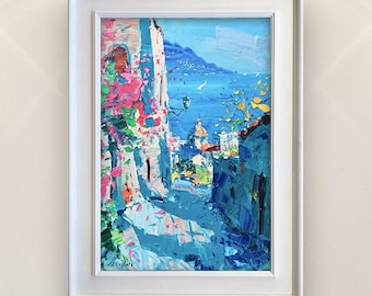 May include: A framed oil painting depicts a vibrant coastal scene. The artwork features a narrow street with buildings in shades of blue, pink, and white, leading to a bright blue sea. The painting is encased in a white frame.