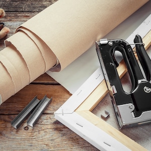 May include: A close-up of a staple gun, staples, and a roll of canvas on a wooden surface. The staple gun is black with a silver handle and is used to attach the canvas to the wooden frame.