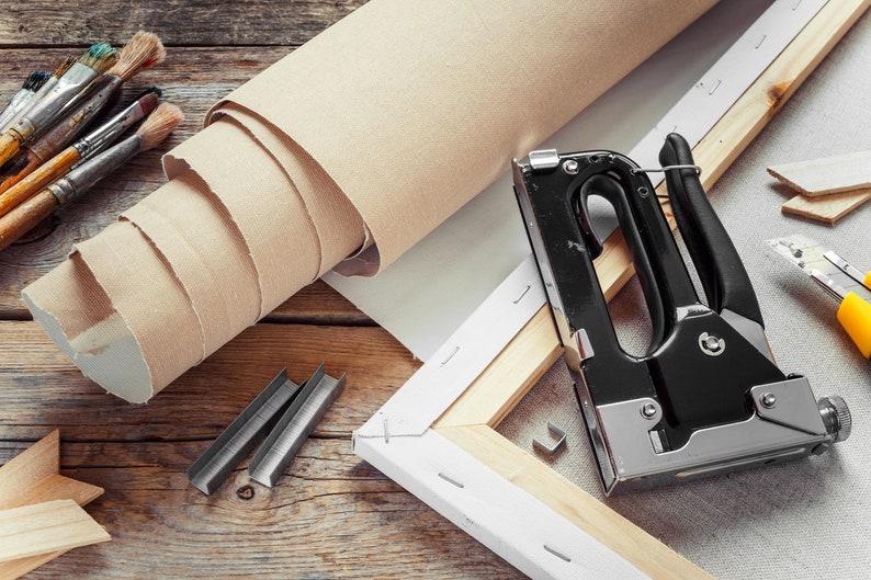 May include: A close-up of a staple gun, staples, and a roll of canvas on a wooden surface. The staple gun is black with a silver handle and is used to attach the canvas to the wooden frame.