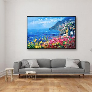 Original hand-painted oil painting on canvas depicting Positano on the Amalfi Coast, with colorful cliffside houses, deep blue sea, and vibrant flowers in the foreground, framed and hanging above a gray sofa in a modern minimalist living room.