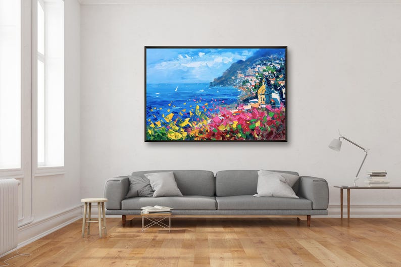 Original hand-painted oil painting on canvas depicting Positano on the Amalfi Coast, with colorful cliffside houses, deep blue sea, and vibrant flowers in the foreground, framed and hanging above a gray sofa in a modern minimalist living room.