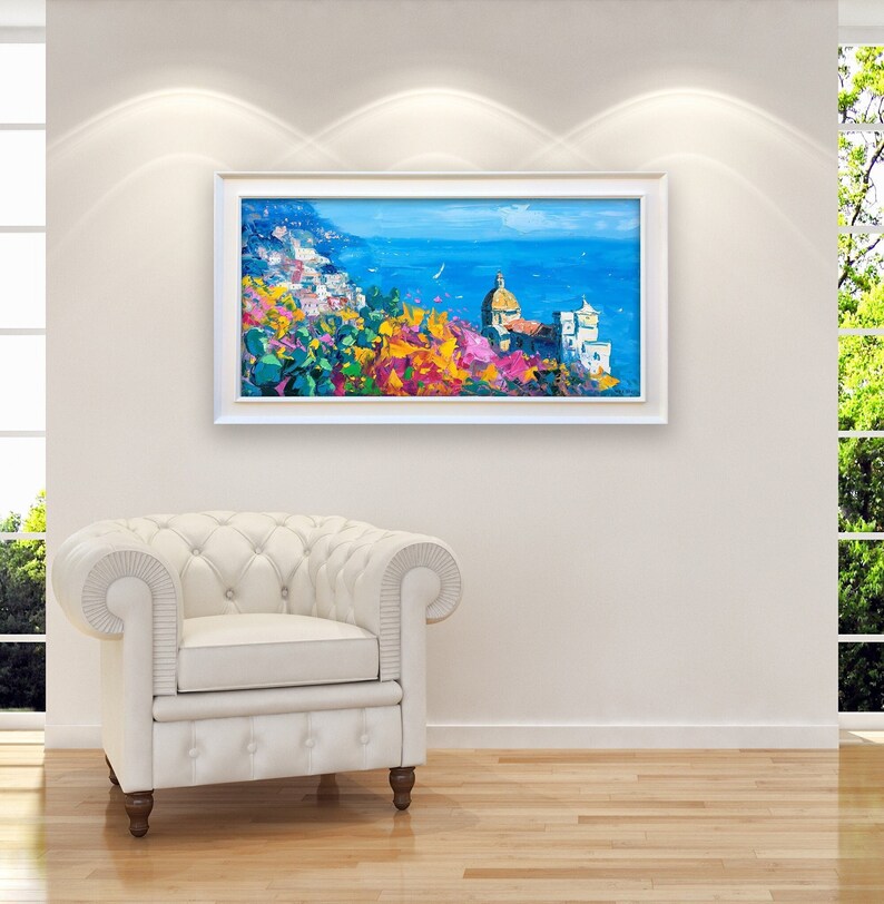 Framed panoramic Positano oil painting with church and floral foreground displayed in a bright living room interior, with a white wooden frame.