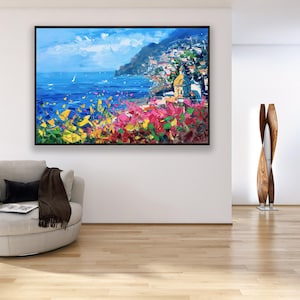 Original hand-painted oil painting on canvas depicting Positano on the Amalfi Coast, with colorful cliffside houses overlooking the deep blue sea and vibrant flowers in the foreground, framed and hanging on a modern living room wall above a light armchair.