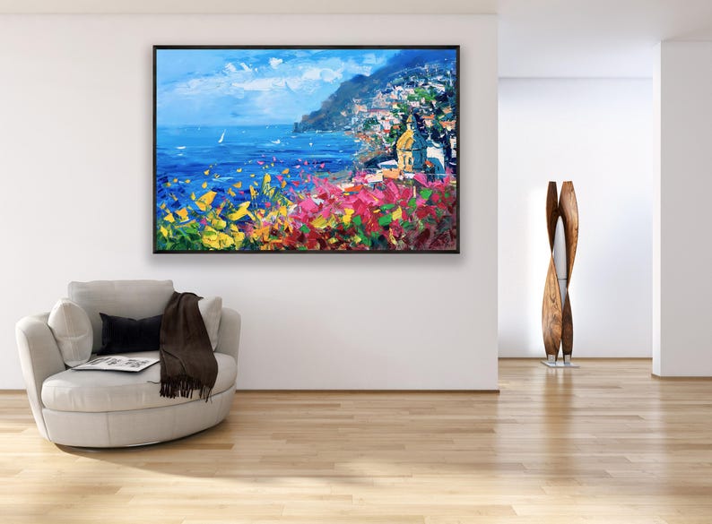 Original hand-painted oil painting on canvas depicting Positano on the Amalfi Coast, with colorful cliffside houses overlooking the deep blue sea and vibrant flowers in the foreground, framed and hanging on a modern living room wall above a light armchair.