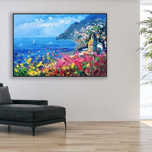 Original hand-painted oil painting on canvas depicting Positano on the Amalfi Coast, with colorful cliffside houses, deep blue sea, and vibrant Mediterranean flowers in the foreground, framed and hanging on a modern living room wall above a black chaise lounge.