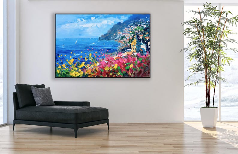 Original hand-painted oil painting on canvas depicting Positano on the Amalfi Coast, with colorful cliffside houses, deep blue sea, and vibrant Mediterranean flowers in the foreground, framed and hanging on a modern living room wall above a black chaise lounge.
