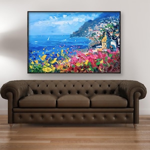 Original hand-painted oil painting on canvas depicting Positano on the Amalfi Coast, with colorful cliffside houses, deep blue sea, and vibrant Mediterranean flowers in the foreground, framed and hanging above a dark sofa in a contemporary living room.