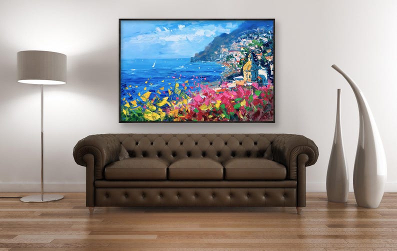 Original hand-painted oil painting on canvas depicting Positano on the Amalfi Coast, with colorful cliffside houses, deep blue sea, and vibrant Mediterranean flowers in the foreground, framed and hanging above a dark sofa in a contemporary living room.