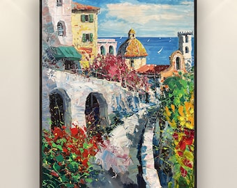 May include: Oil painting of a coastal town scene with buildings in shades of white, yellow, and orange. The artwork features a blue sky, sea, and colorful flowers. The painting is framed in a black border.