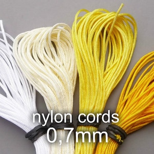 Puede incluir: Tres carretes de cordón de nylon en blanco, crema y amarillo. El cordón tiene un grosor de 0,7 mm. Los carretes están etiquetados con "nylon cords 0,7 mm".