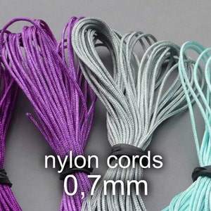 Puede incluir: Tres carretes de cordón de nylon en morado, gris y azul claro. Los cordones tienen un grosor de 0,7 mm y están agrupados. El texto "nylon cords 0,7mm" está impreso en la imagen.