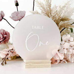 May include: White frosted acrylic round table number with the text "TABLE One" on a wooden stand. The table number is surrounded by pink and white flowers.