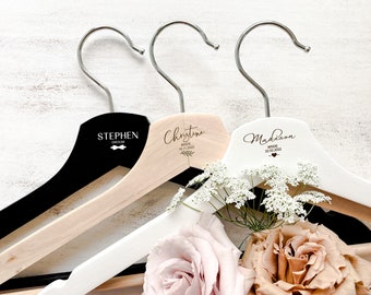 May include: Three wooden hangers with personalized names and wedding dates. The hangers are in black, natural wood, and white. The hangers are decorated with a bow tie, a floral design, and a heart.