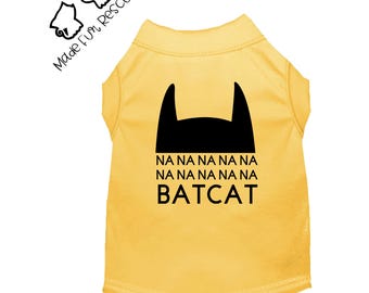 May include: A yellow sleeveless pet shirt featuring a black graphic of a bat-like mask silhouette above the text NA NA NA NA NA NA NA NA NA BATCAT. The logo Made Fur Rescues is printed in black in the upper left corner of the image.