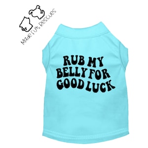 Puede incluir: Una camiseta para mascota de color azul claro con el texto negro "RUB MY BELLY FOR GOOD LUCK" en una fuente ondulada y negrita. La prenda incluye un logotipo en la esquina superior izquierda con el texto "Made Fur Rescues" y dos cabezas de perro.