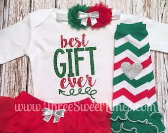 Puede incluir: Un conjunto de Navidad para niña con un body blanco que dice "best gift ever" en purpurina verde y roja, un tutú rojo de tul con un lazo plateado, polainas a rayas verdes y rojas con un corazón plateado y una diadema de flores verdes y rojas con un lazo plateado.