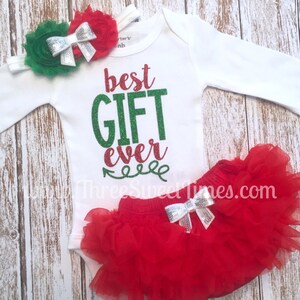 Puede incluir: Un body blanco de manga larga con el texto "best GIFT ever" en purpurina verde y roja. El body se combina con una falda de tul roja y una diadema de flores verde y roja con un lazo plateado.